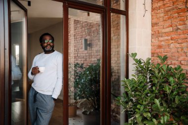 Black bearded man drinking coffee while standing in doorway at home