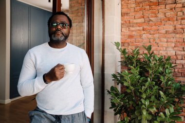 Black bearded man drinking coffee while standing in doorway at home