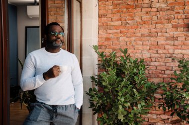 Black bearded man drinking coffee while standing in doorway at home