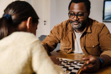 Black girl and her father playing checkers in kitchen at home