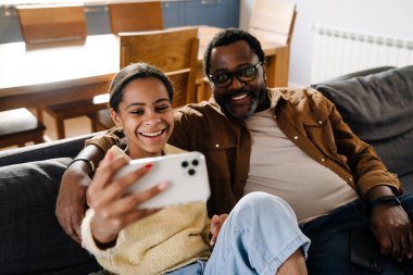 Black girl and her father taking selfie on cellphone while sitting on sofa at home