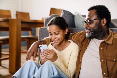Black girl and her father hugging and using cellphone while sitting on floor at home