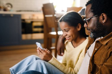 Black girl and her father hugging and using cellphone while sitting on floor at home