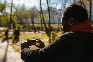 Black bearded man taking photo on cellphone while strolling in park outdoors