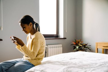 Black brunette girl using mobile phone while sitting on bed at home
