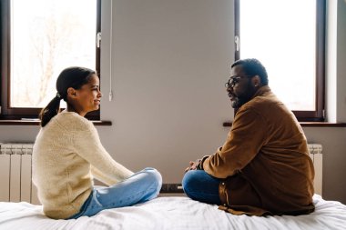 Black girl and her father talking while sitting on bed at home