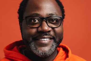 Black bearded man in eyeglasses smiling and looking at camera isolated over orange background