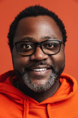 Black bearded man in eyeglasses smiling and looking at camera isolated over orange background