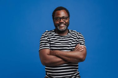 Black bearded man in eyeglasses smiling and looking at camera isolated over blue background
