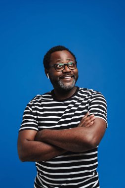 Black man in eyeglasses smiling while listening music with earphones isolated over blue background