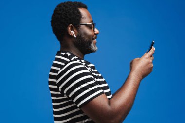 Black man in earphones listening music while using mobile phone isolated over blue background