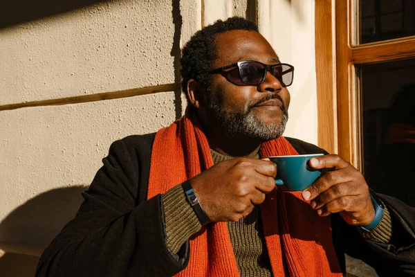 Black bearded man wearing sunglasses drinking coffee while sitting in street cafe outdoors