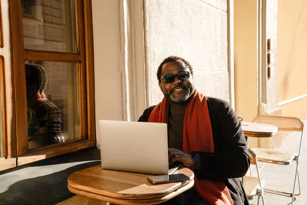 Black bearded man using laptop while sitting in cafe outdoors