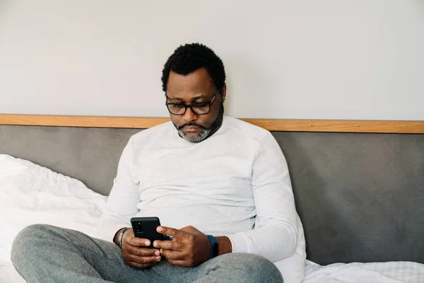 Black Man Wearing Eyeglasses Using Mobile Phone While Resting Bed — Stockfoto
