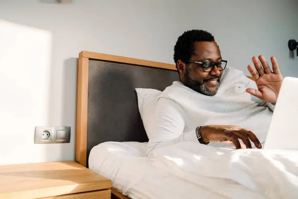 Black man waving hand and using laptop while resting in bed at home