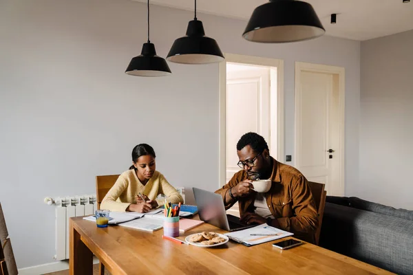 Black man working with laptop while his daughter doing homework at home