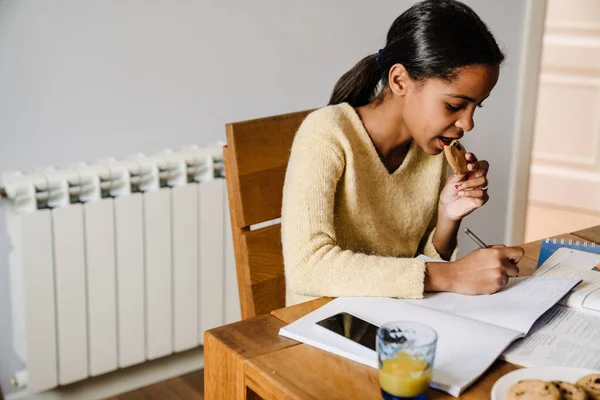 Black Happy Girl Eating Cookie While Doing Homework Kitchen Home — Stok fotoğraf