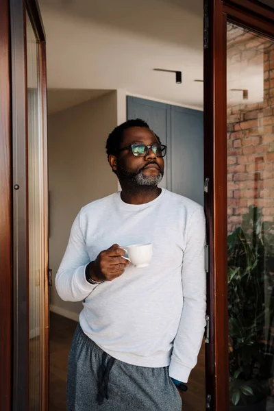 Black bearded man drinking coffee while standing in doorway at home
