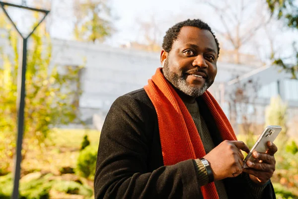 Black bearded man smiling and using cellphone while strolling in park