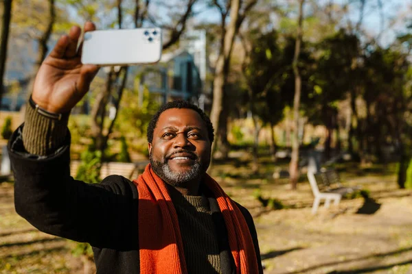 Black bearded man taking selfie on cellphone while strolling in park outdoors