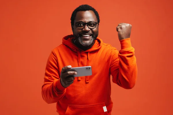 Black bearded man making winner gesture and using cellphone isolated over orange background