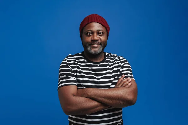 Black bearded man wearing hat smiling and looking at camera isolated over blue background