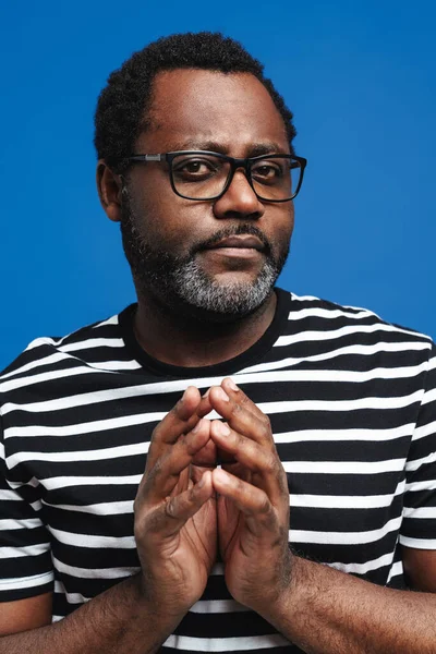 Black bearded man in eyeglasses gesturing and looking at camera isolated over blue background