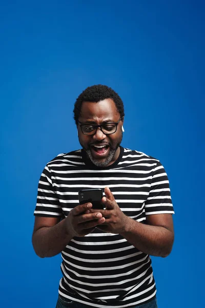 Black man in earphones listening music while using mobile phone isolated over blue background