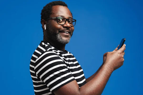 Black man in earphones listening music while using mobile phone isolated over blue background