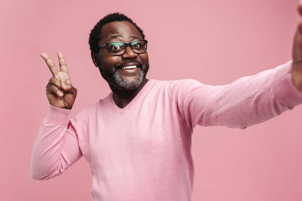 Black bearded man in eyeglasses gesturing while taking selfie photo isolated over pink background