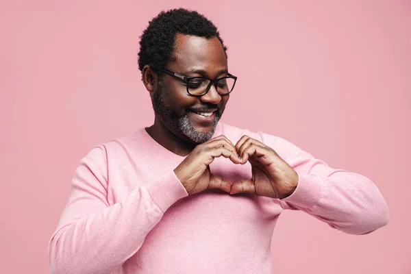 Black bearded man in eyeglasses smiling and making heart gesture isolated over pink background