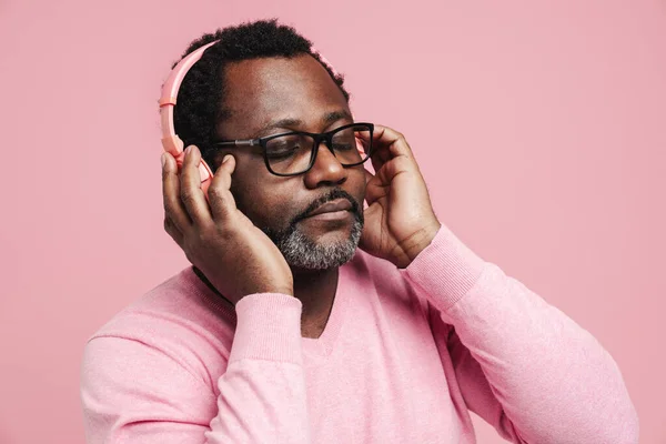 Black bearded man in eyeglasses listening music with headphones isolated over pink background
