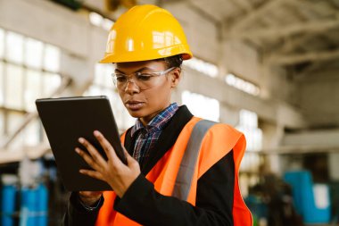 Black woman wearing helmet and vest working with tablet computer at factory