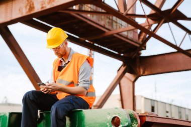 White ginger man wearing helmet and vest using cellphone working in port