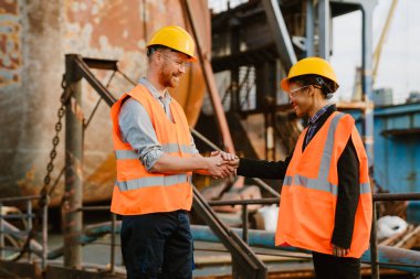 Multiracial man and woman wearing helmets handshaking while working at port