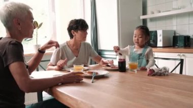 Positive mature lesbian couple eating breakfast with adopted Asian daughter in the kitchen at home