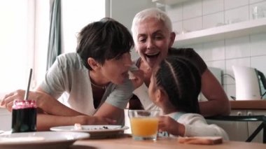 Cheerful mature lesbian couple eating breakfast with adopted Asian daughter in the kitchen at home