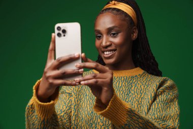 Black young woman with pigtails taking selfie on cellphone isolated over green wall