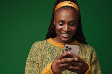 Black young woman with pigtails smiling while using cellphone isolated over green wall