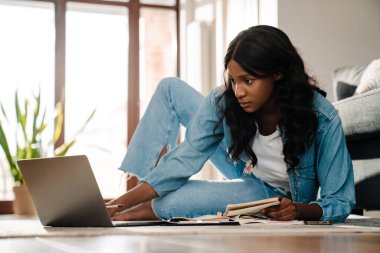 Black young woman studying and using laptop while sitting on floor at home