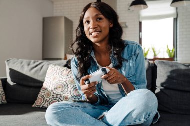 Black young woman smiling while playing video games and sitting on couch at home