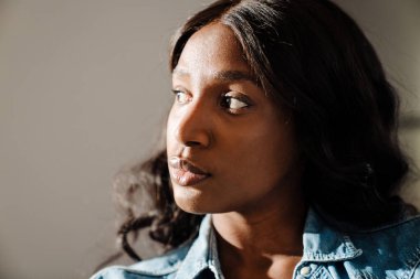 Black young woman wearing denim shirt looking aside while sitting over beige wall