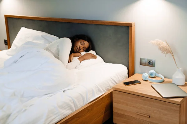 Black young woman sleeping while lying on white bed linen at home