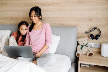 Asian mother and daughter using laptop while sitting on bed at home