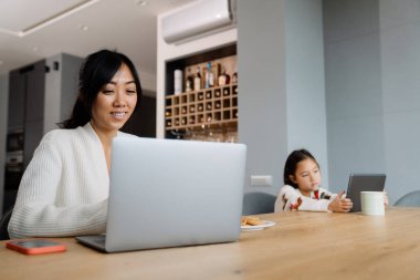 Asian woman and her daughter using gadgets while sitting at table in home