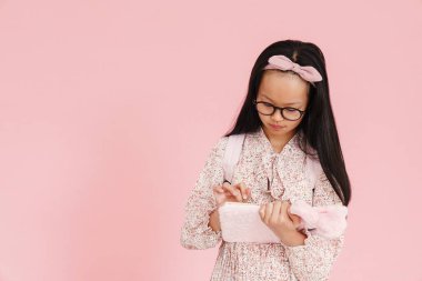 Asian girl wearing eyeglasses writing down notes in exercise book isolated over pink background
