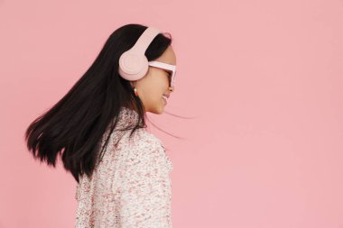 Asian girl dancing while listening music with headphones isolated over pink background