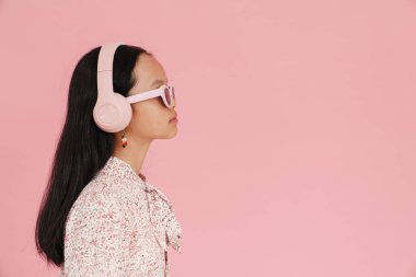 Asian girl wearing sunglasses listening music with headphones isolated over pink background