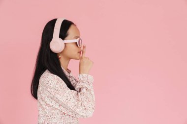 Asian girl listening music while showing silence gesture isolated over pink background