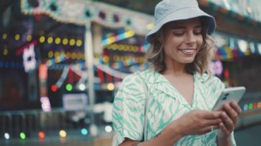 Positive young woman wearing panama hat typing by phone and looking at the camera while standing in adventure park